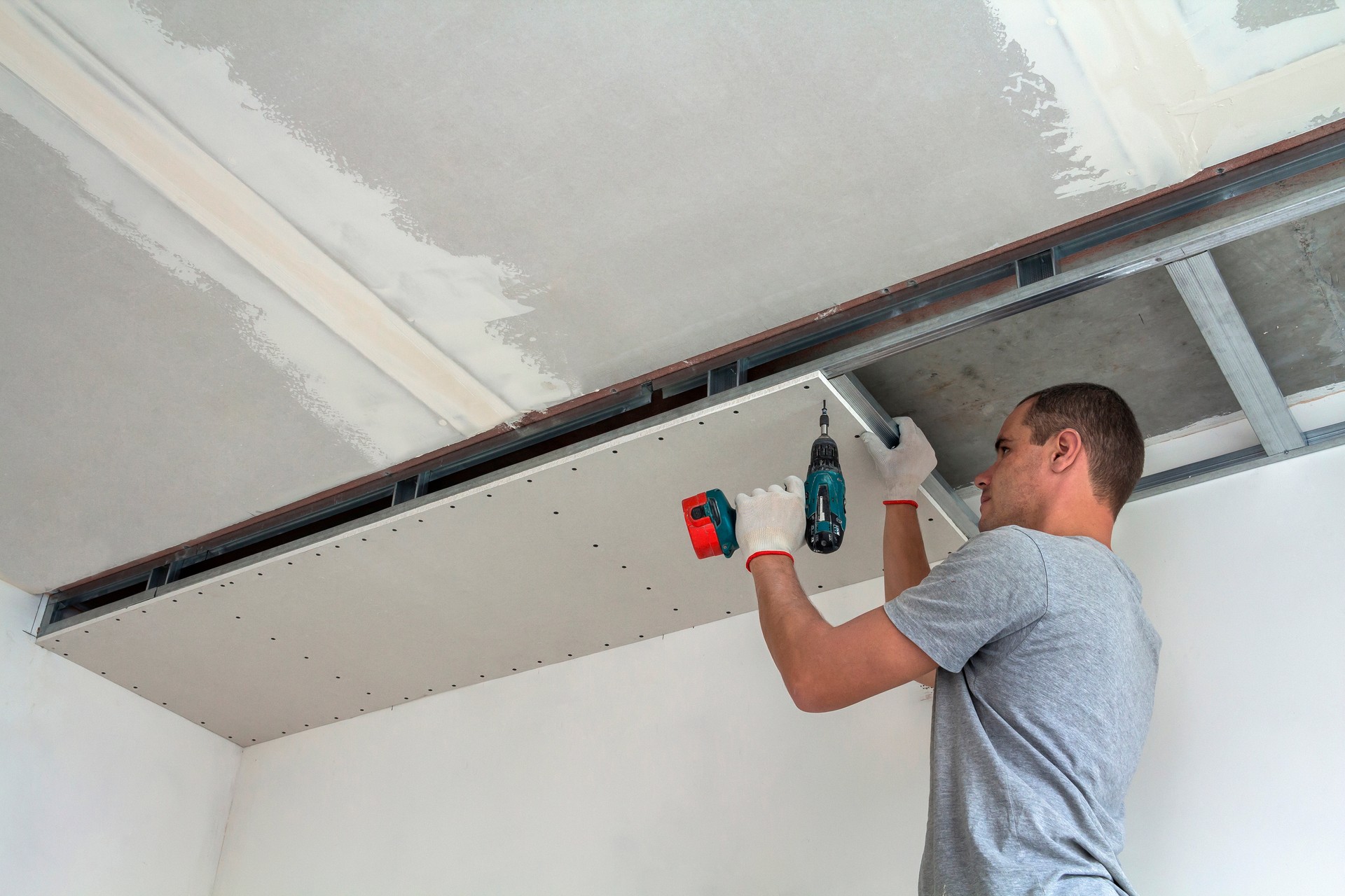 Young man in usual clothing and work gloves fixing drywall suspended ceiling to metal frame using electrical screwdriver on ceiling insulated with shiny aluminum foil. DIY, do it yourself concept.